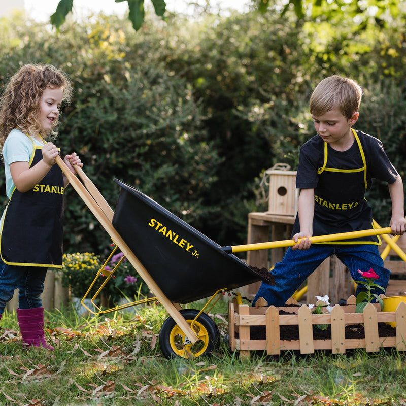 Children using Stanley Jr black wheelbarrow with yellow handles in garden setting