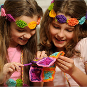 Two young girls smiling and crocheting together, each wearing a colorful flower headband. They are holding a handmade multicolor crochet granny square piece with a purple crochet hook.
