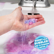 Hand holding colorful mini bath bombs under running water creating purple bubbles