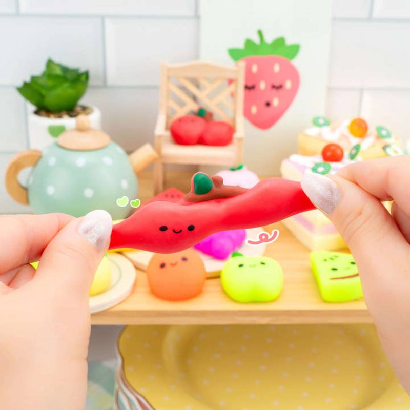 Hands holding a red chili pepper mochi squishy among assorted kawaii fruit squishies on wooden tray