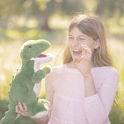 Young woman holding green T-Rex dinosaur plush toy outdoors in sunlight
