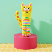 Yellow paper mache cat with colorful stripes on pink pedestal against mint background