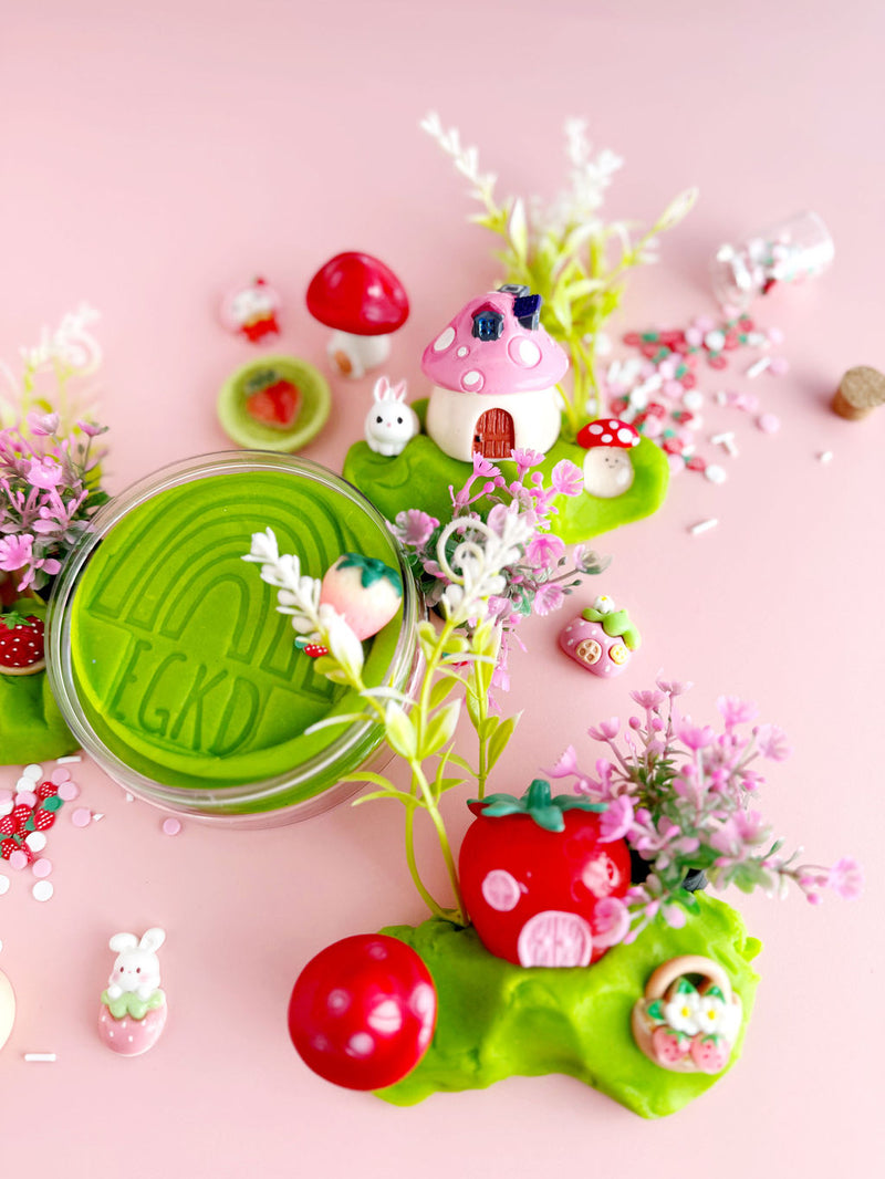 Colorful playdough village scene with mushroom houses, bunny figures, flowers on pink background
