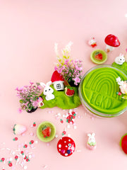 Green play dough with cute bunny figurines, miniature houses, and strawberry decorations on pink background