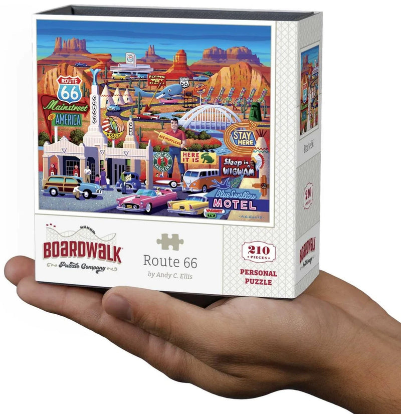 A hand holds a white puzzle box featuring a colorful illustrated desert motel scene with Route 66 signs, cars, and desert rock formations. The box reads Boardwalk, Route 66, by Andy C.