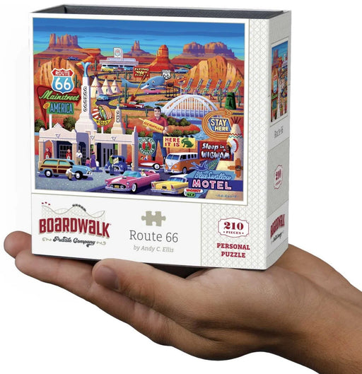 A hand holds a white puzzle box featuring a colorful illustrated desert motel scene with Route 66 signs, cars, and desert rock formations. The box reads Boardwalk, Route 66, by Andy C.