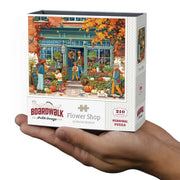 A hand holding a boxed jigsaw puzzle. The front of the box shows an illustrated autumn flower shop storefront with people, pumpkins, flowers, and fall foliage.
