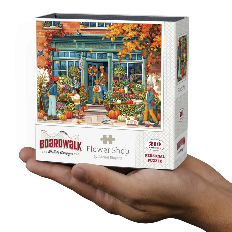 A hand holding a boxed jigsaw puzzle. The front of the box shows an illustrated autumn flower shop storefront with people, pumpkins, flowers, and fall foliage.