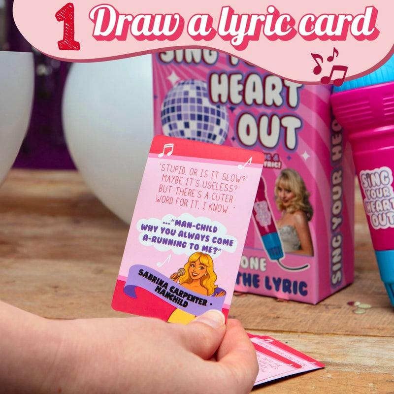 A hand holds a pink lyric card from the 'Sing Your Heart Out' game, with the product box and matching pink cup in the background on a wooden table. A pink promotional banner at the top reads '1 Draw a lyric card'.