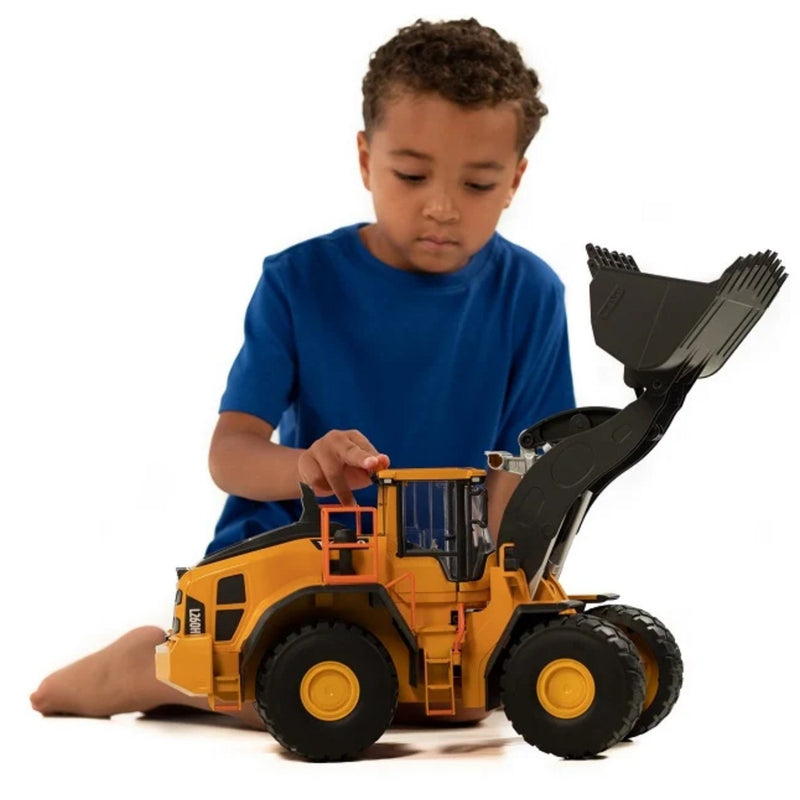 A young boy in a blue t-shirt sits on the floor playing with a large yellow and black toy wheel loader construction vehicle against a white background.