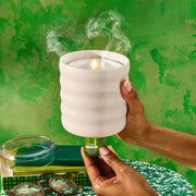 Hands holding cream-colored flameless candle diffuser with steam rising against green background