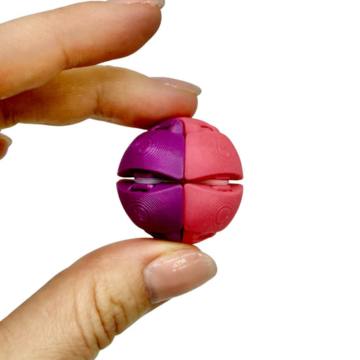 A hand holding a small spherical puzzle toy with four interlocking pink and purple sections on a plain white background.