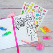 A styled flat-lay of a unicorn-themed notebook with the words 'BE Magical!' and a black-and-white unicorn illustration, surrounded by neon highlighters, sticker sheets, and a pink pom-pom pen on a whitewashed wood surface.