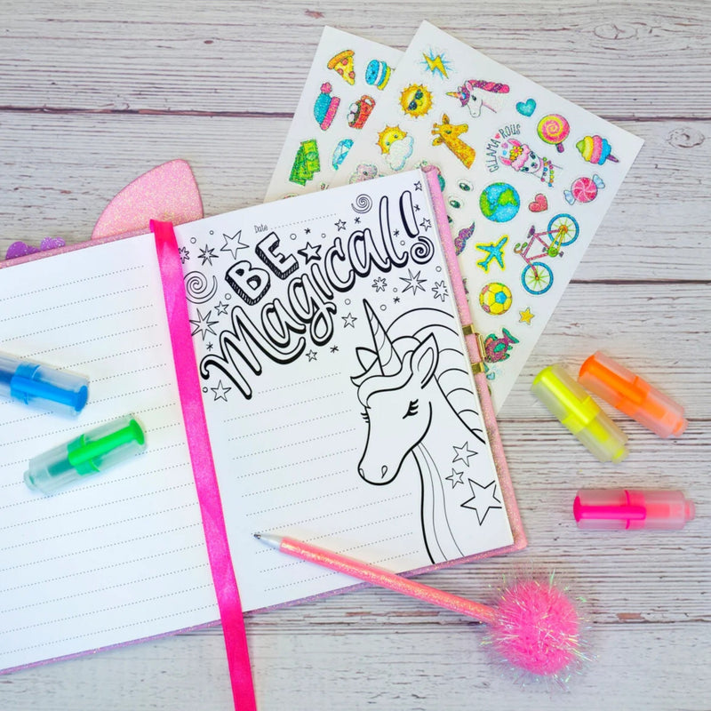 A styled flat-lay of a unicorn-themed notebook with the words 'BE Magical!' and a black-and-white unicorn illustration, surrounded by neon highlighters, sticker sheets, and a pink pom-pom pen on a whitewashed wood surface.