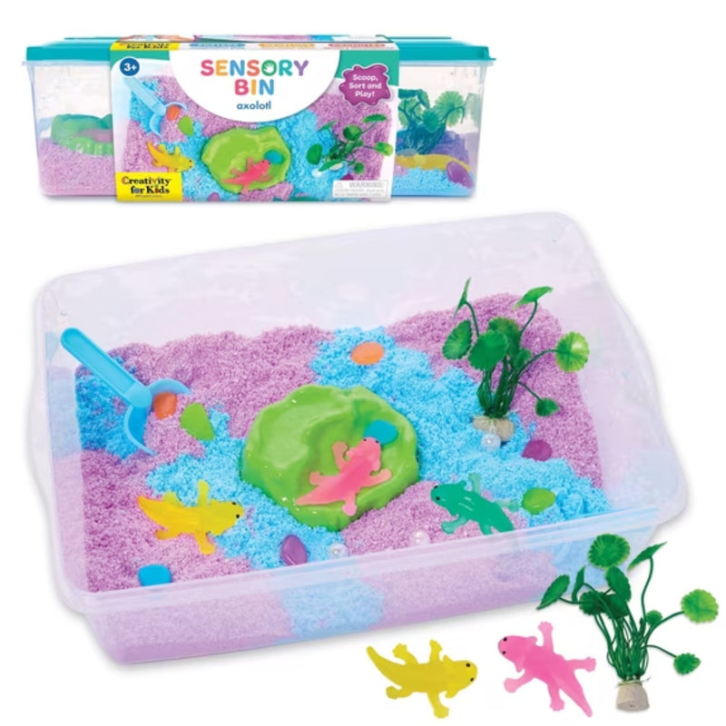 A children's sensory bin set shown open with pink and blue sand, toy axolotls, a green pond, plastic plants, and small animal figures, plus the product box in the background. The set is photographed on a clean white studio background.