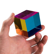 A hand holding a metallic, iridescent cube puzzle toy that reflects multiple colors including blue, yellow, orange, purple, and dark blue against a plain white background.