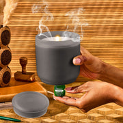 Frosted charcoal flameless candle diffuser with hands holding essential oil bottle in spa setting