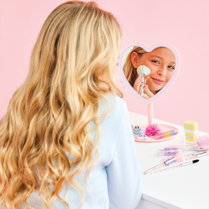 Young girl using heart-shaped vanity mirror with makeup brush in pink bedroom setting