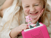 Young girl smiling while playing with pink Tonie Care Bears Cheer Bear on Toniebox