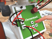 A close-up of a Clementoni robotics control panel with a green faceplate, white housing, red and black wires, and a finger pressing one of the square buttons.