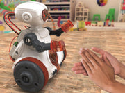 A small white robot toy with black and red wheels and translucent orange arms is shown on a wooden floor in a toy-filled room, with a person's hands reaching toward it.