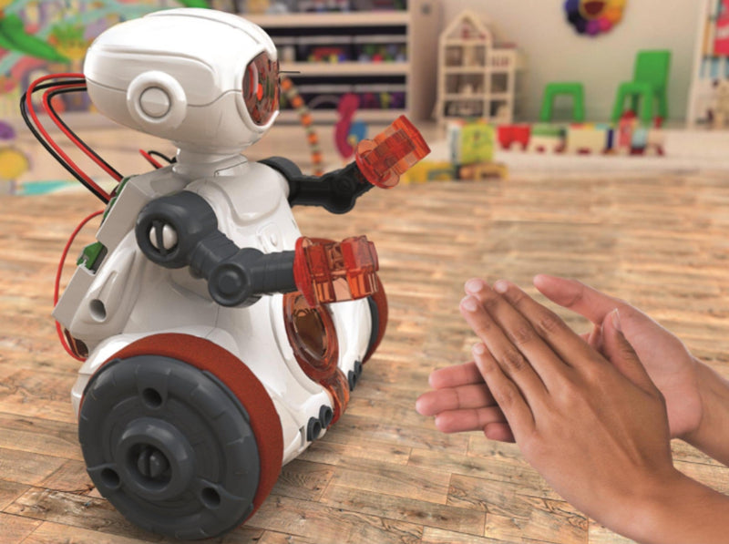 A small white robot toy with black and red wheels and translucent orange arms is shown on a wooden floor in a toy-filled room, with a person's hands reaching toward it.