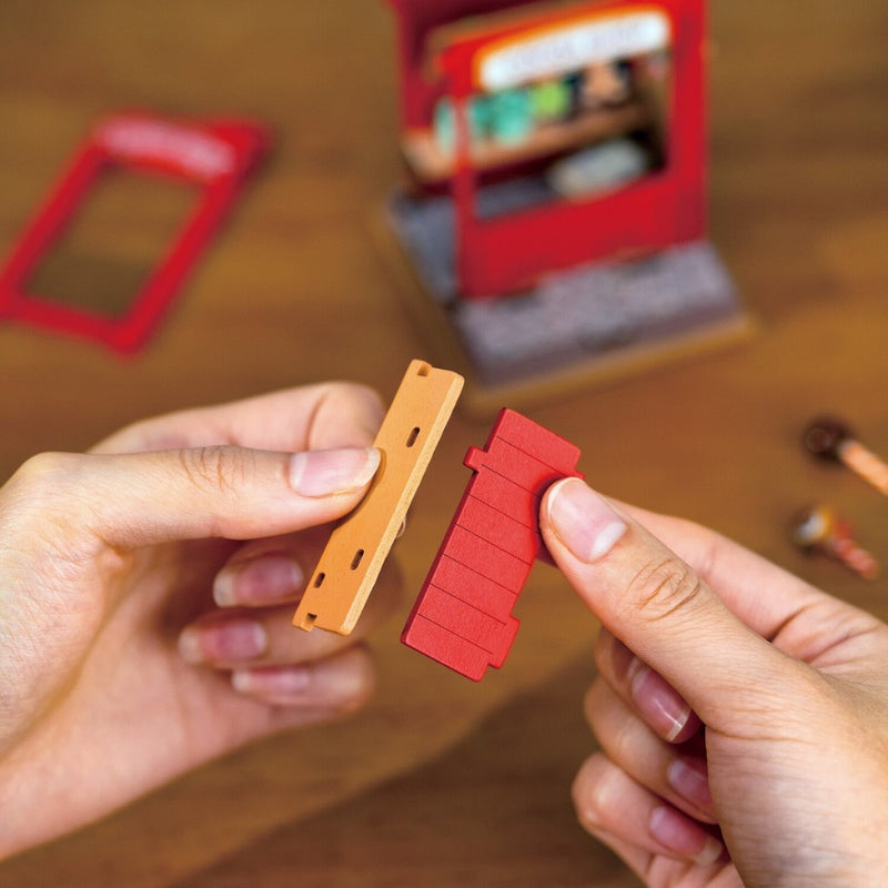 Two hands holding a red and orange plastic model piece on a wooden surface with blurred toy store in the background.