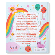 A children's book back cover shown flat against a white background. The cover features a light blue base with a large rainbow, smiling sunflower, balloons, stars, clouds, and a pink cat, surrounding a central text panel with a message about being grateful and big-hearted.