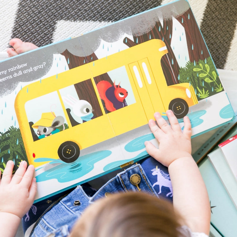 A baby's hands and head are visible as they hold and explore a colorful children's book featuring a yellow school bus with cartoon animals in a rainy forest scene.