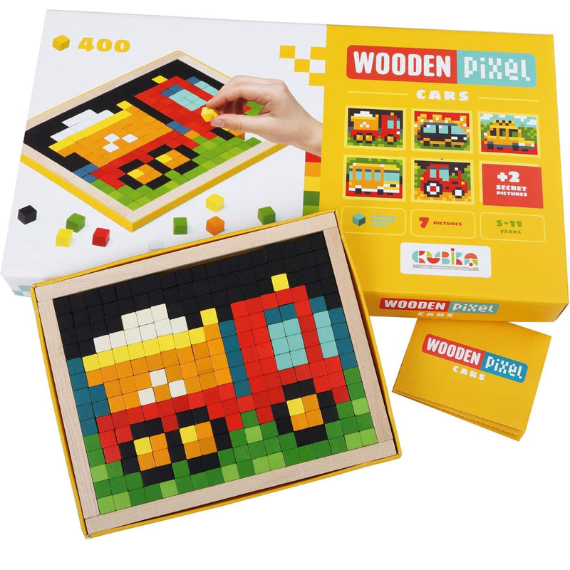 A boxed toy set labeled 'Wooden Pixel Cars' shown with its packaging, a completed pixel-art car board, and a small card. A hand is visible placing a piece on the box, so by the schema this is Lifestyle rather than Product/Packaging, even though the scene is still product-focused.