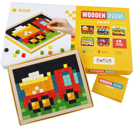 A boxed toy set labeled 'Wooden Pixel Cars' shown with its packaging, a completed pixel-art car board, and a small card. A hand is visible placing a piece on the box, so by the schema this is Lifestyle rather than Product/Packaging, even though the scene is still product-focused.