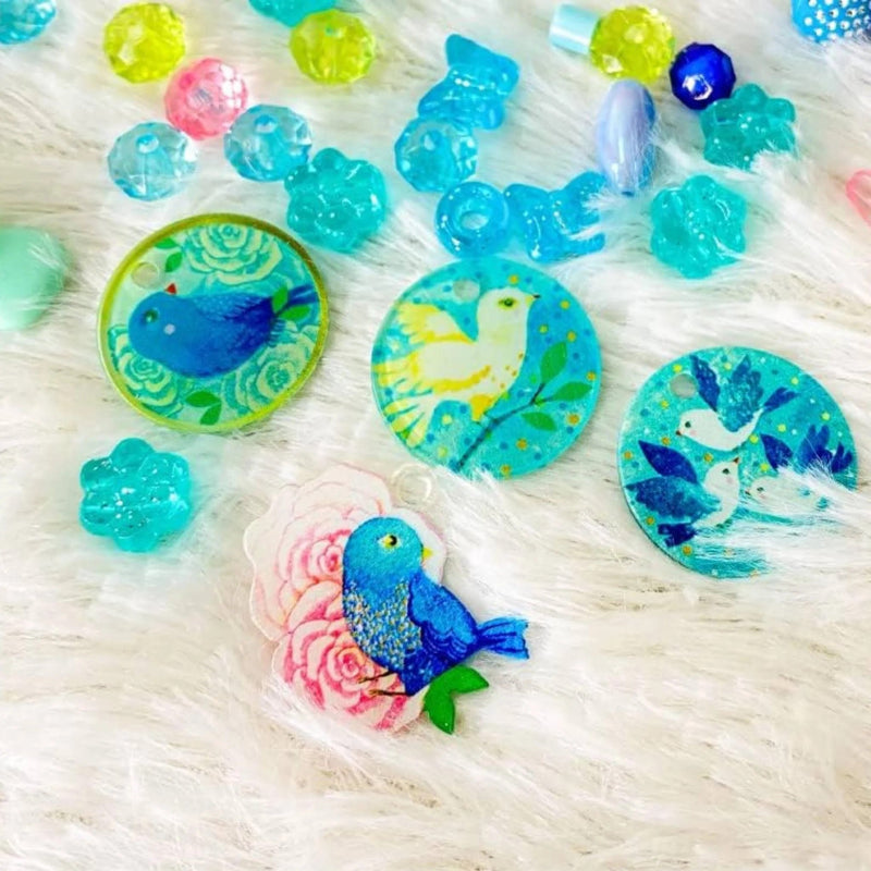 A collection of colorful handmade glass beads and pendants featuring painted blue birds, white doves, and floral designs, arranged on a white faux fur background.