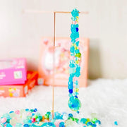 A colorful beaded hanging ornament or charm made of translucent blue, turquoise, green, and pink beads, suspended from a thin metal stand on a white surface with blurred pink boxes in the background.