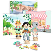 A children's play set featuring three animal doll figures (a fox, a dog, and a cat) on wooden bases, with colorful clothing accessories and a fold-out backdrop showing a Parisian bridge and a pink bakery storefront.