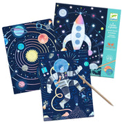 A set of three colorful space-themed glitter art cards on a white background, featuring illustrations of planets, a rocket ship, and an astronaut. A wooden stick is placed diagonally across the cards, and the packaging box is visible on the right.