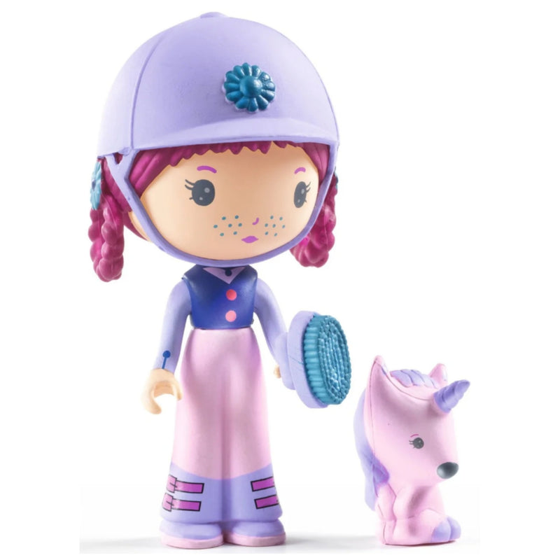 A stylized doll figure wearing a lavender equestrian helmet and matching outfit, holding a blue grooming brush, with a small pink-and-lavender unicorn toy beside it on a white background.