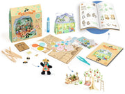 Djeco's wooden building and craft set arranged on a white background, including a colorful box, small house kit, crayon, instruction booklet, wooden pieces, pegs, paint palette, tweezers, and assembled toy structures.