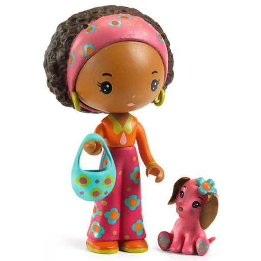 A colorful toy doll with dark curly hair, a pink patterned headband, an orange top, and pink patterned pants, holding a turquoise patterned handbag, standing next to a small pink dog figurine on a white background.