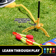 A yellow plastic toy water fountain stand on grass with several red-and-yellow foam water rockets nearby. Text on the image says 'EXPERIMENT WITH ANGLES, TRAJECTORY AND FORCE' and a banner at the bottom reads 'LEARN THROUGH PLAY' with STEM icons.