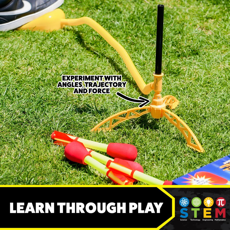 A yellow plastic toy water fountain stand on grass with several red-and-yellow foam water rockets nearby. Text on the image says 'EXPERIMENT WITH ANGLES, TRAJECTORY AND FORCE' and a banner at the bottom reads 'LEARN THROUGH PLAY' with STEM icons.