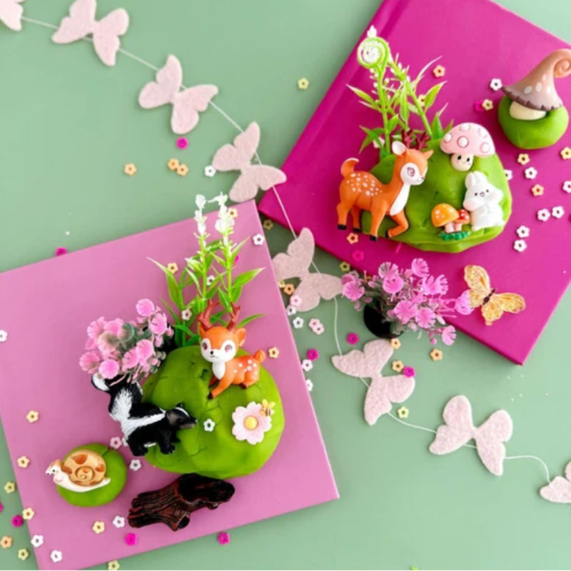 Two pink square craft boards decorated with miniature forest-themed felt pieces, including a deer, mushroom, flower, butterfly, and small animal figures, arranged on a mint green background with scattered felt butterflies and flowers.