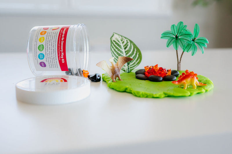 Green playdough creation with palm tree, dinosaur, and campfire scene next to EGKD jar