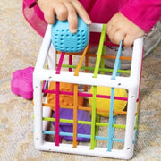 A child's hands holding a blue textured ball over Fat Brain's InnyBin with colorful elastic cords and balls inside, on a carpeted floor.
