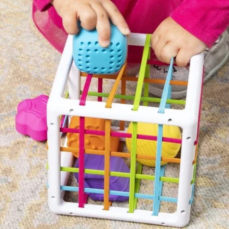 A child's hands holding a blue textured ball over Fat Brain's InnyBin with colorful elastic cords and balls inside, on a carpeted floor.