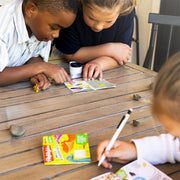 Two children leaning over a wooden table, looking at a small booklet or book. A third child is partially visible in the foreground writing on a page in . Little Explorers activity booklet.