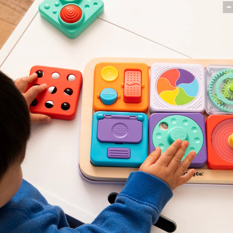 A child's hands interacting with a colorful PlayTab wooden sensory toy board on a white table, with part of the child's head and arm visible.