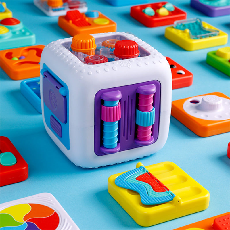 A white plastic fidget activity cube toy with colorful purple, blue, red, and orange buttons and textured panels, shown on a bright blue surface surrounded by other matching colorful toys.
