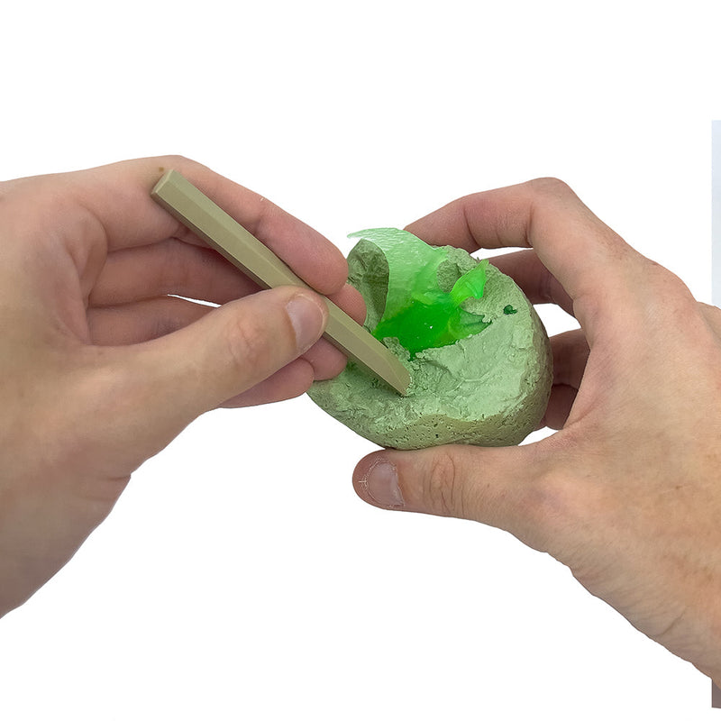 Hands using tool to excavate glowing green dinosaur from egg-shaped fossil kit