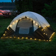 Illuminated tent with warm white globe string lights in lakeside camping setting at dusk