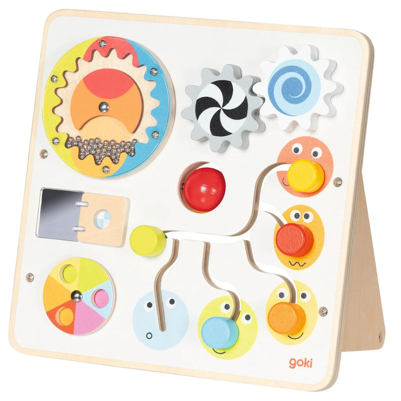 A Goki wooden activity board toy with colorful dials, gears, a sliding bar, and smiling face cutouts, photographed on a clean white background.